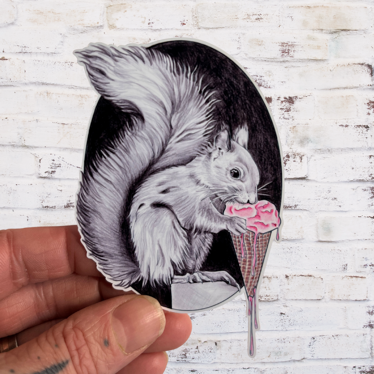 Sticker of a squirrel eating ice cream held against a white brick wall.