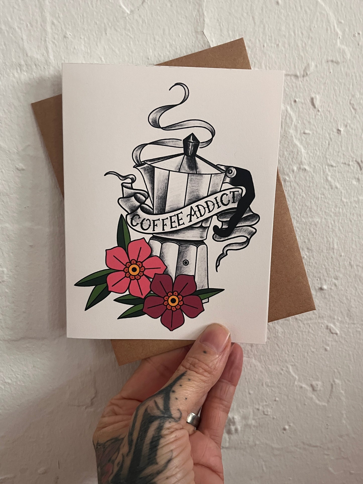 Hand holding a card with tattoo-style design and flowers on a textured white surface