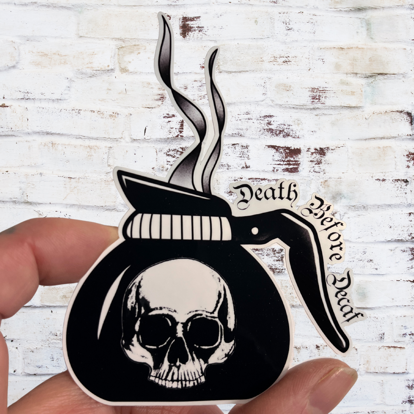 Sticker with skull and coffee pot design and 'Death Before Decaf' text held by a hand against a white brick background