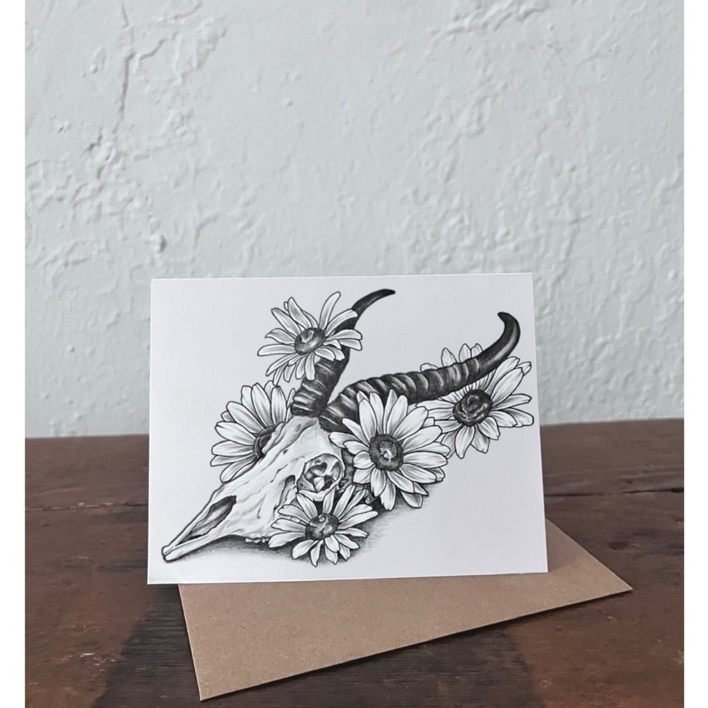 Greeting card with black and white illustration of a springbok skull and daisy flowers on a wooden surface.
