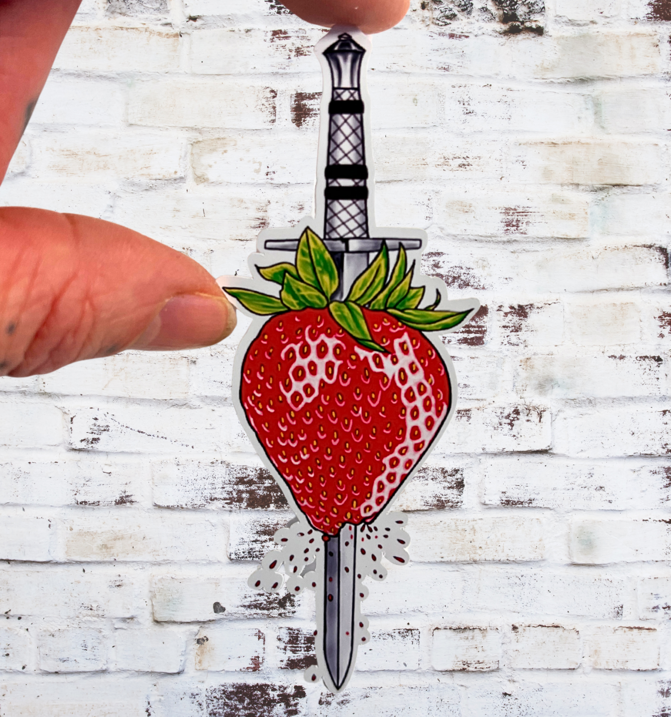 Sticker of a strawberry with a knife through it held by a hand on a white wooden background