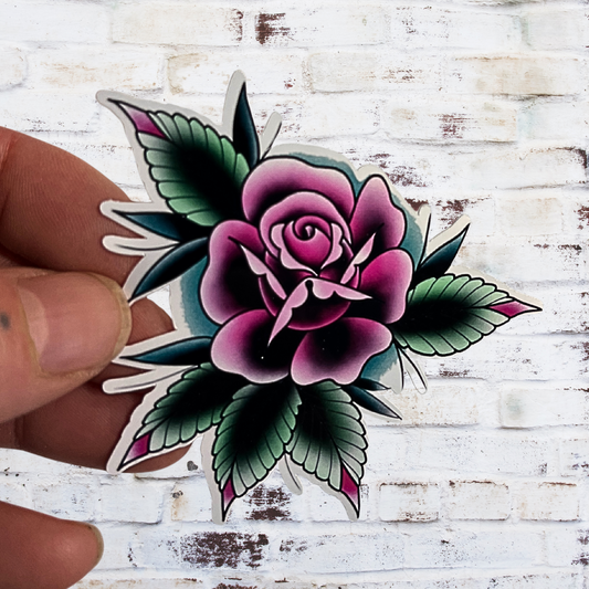 Sticker of a pink rose with green leaves held by a hand against a white wooden background