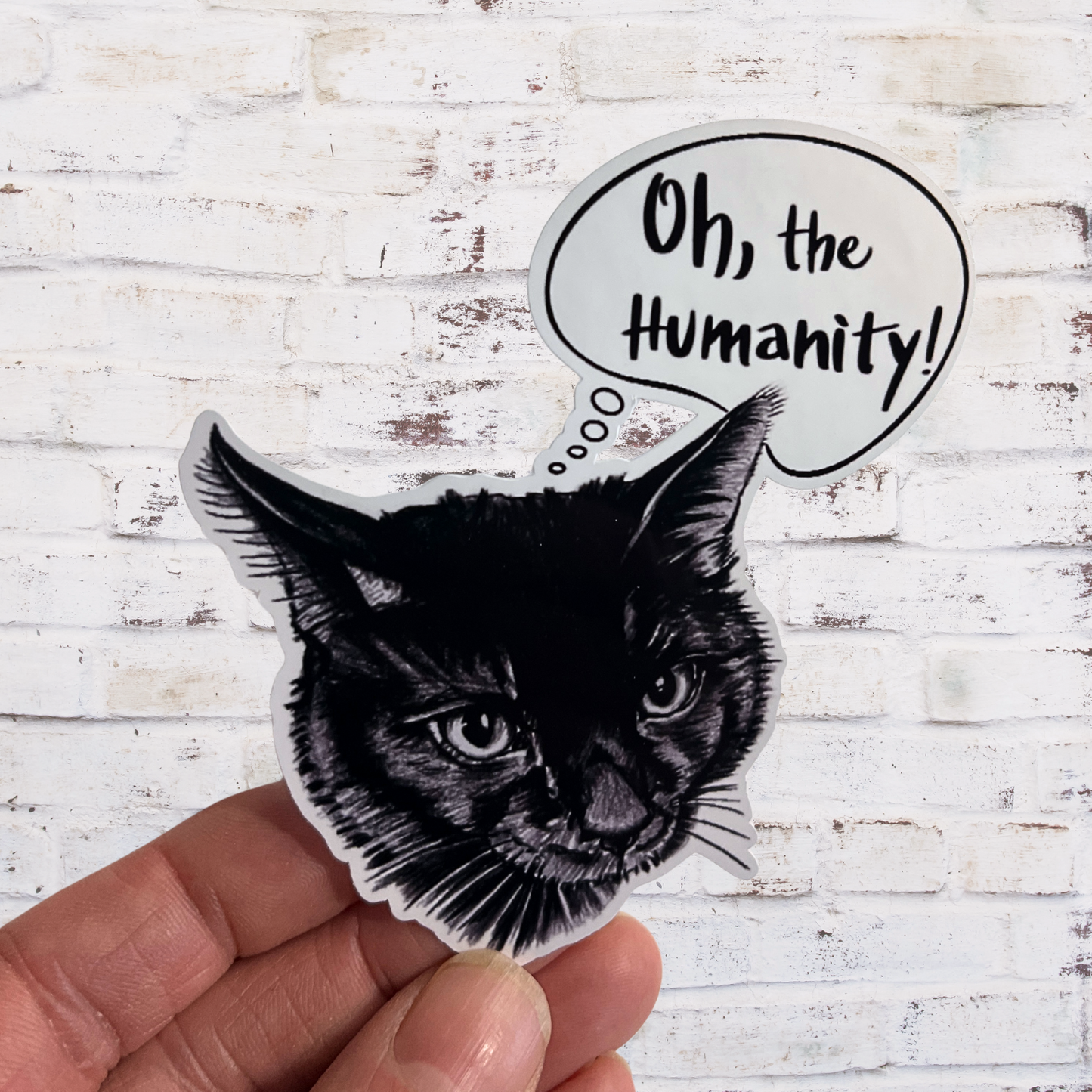 Black and white cat sticker with 'Oh, the Humanity!' text on a white wooden background