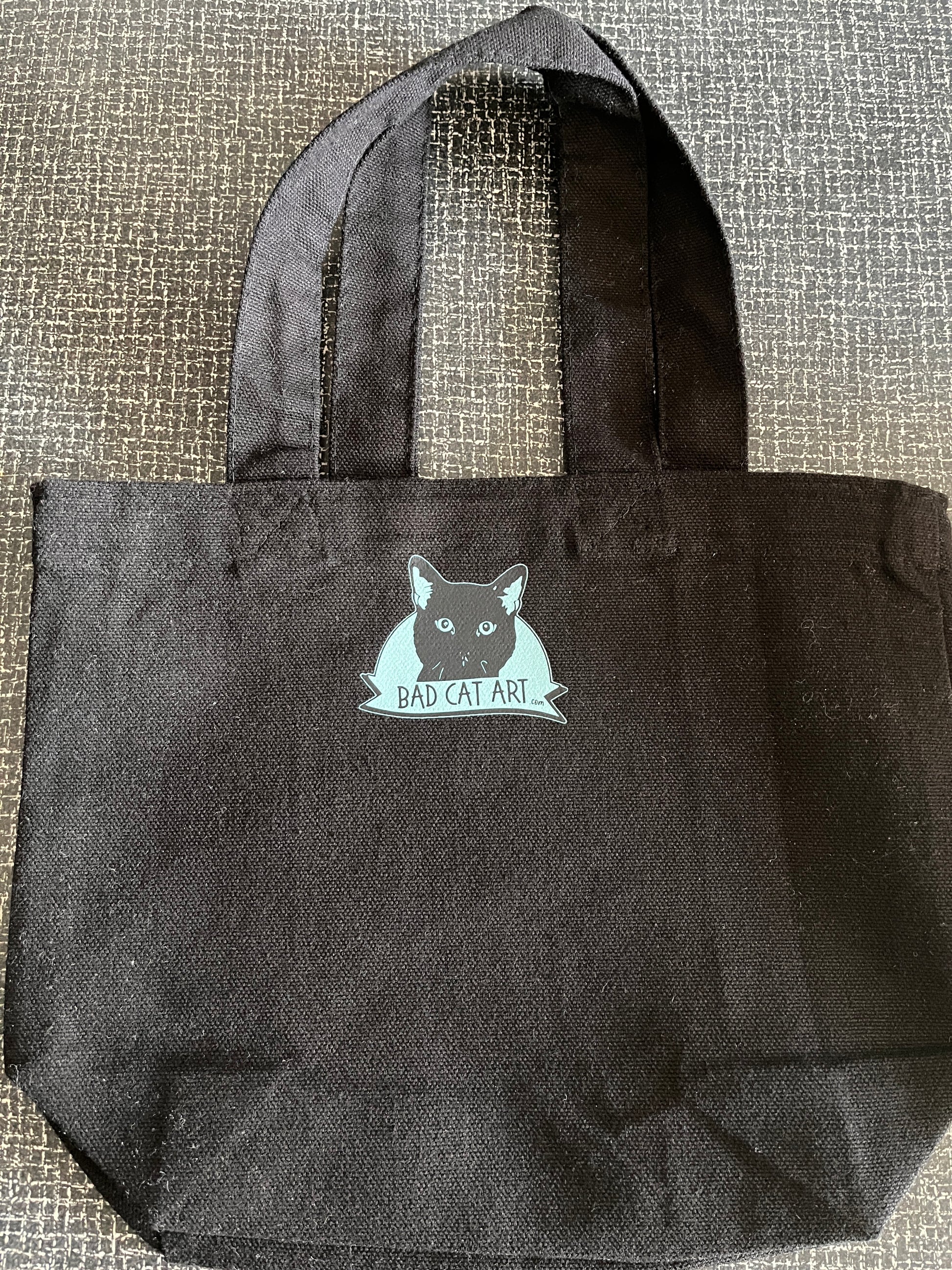 Black tote bag with a cat graphic and text on a textured surface