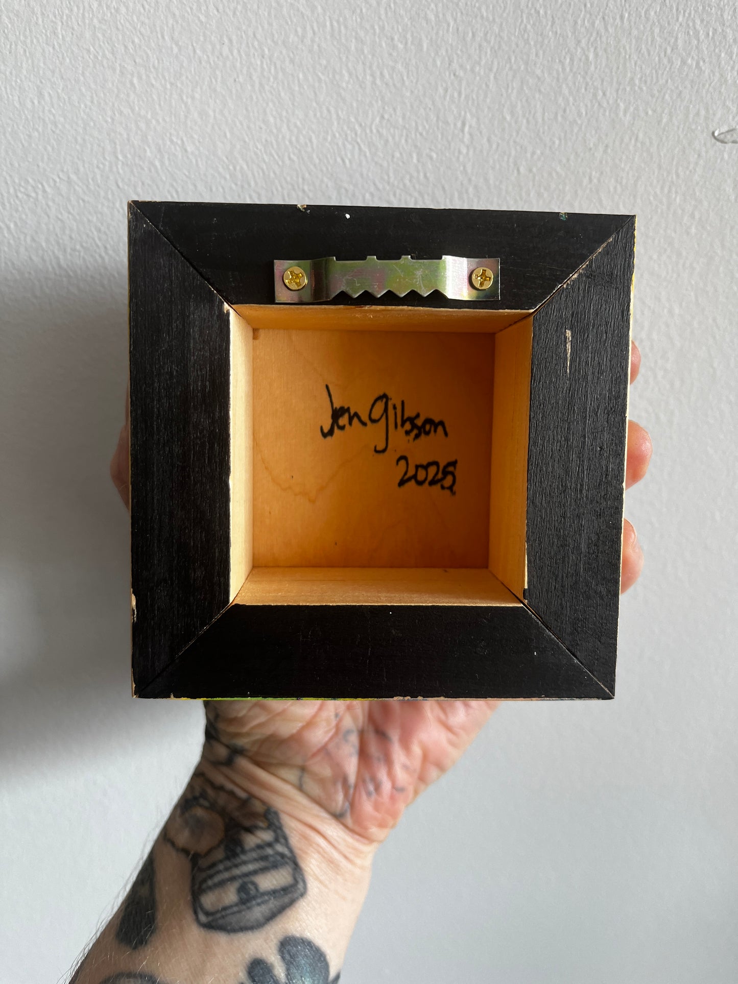 Hand holding a small black wooden box with 'Jon Gibson 2023' written on it against a gray background.