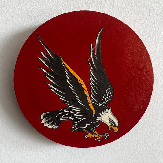 Round painting of a traditional tattoo style eagle design on a red background against a white wall