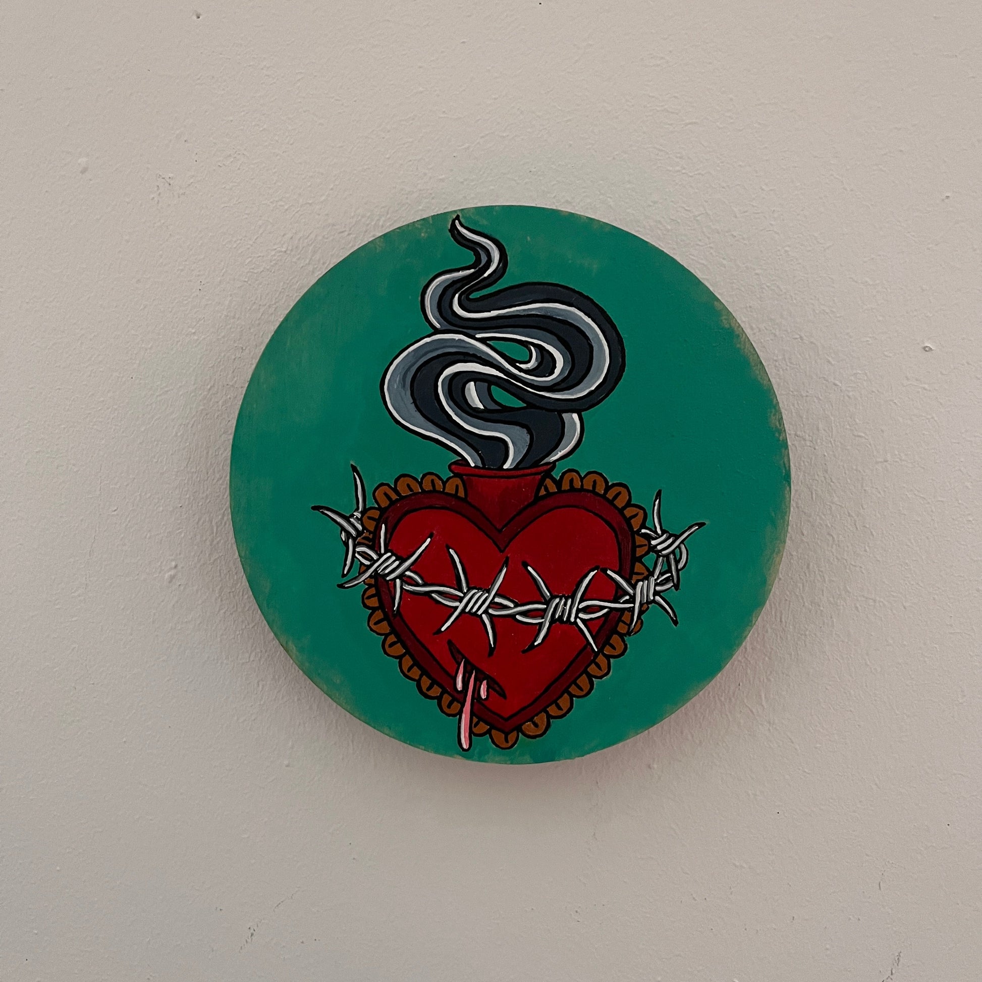 Round teal painted wood panel with a flaming red sacred heart design wrapped with barbed wire and dripping pink blood. 