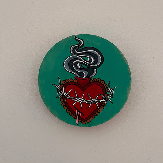 Round teal painted wood panel with a flaming red sacred heart design wrapped with barbed wire and dripping pink blood. 