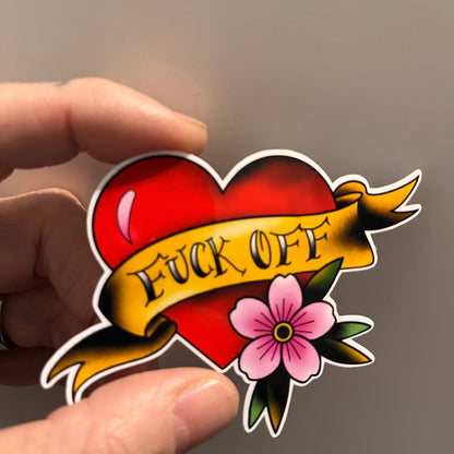 Sticker with red heart and yellow banner with 'F*ck Off', and pink flower held between fingers against a neutral background