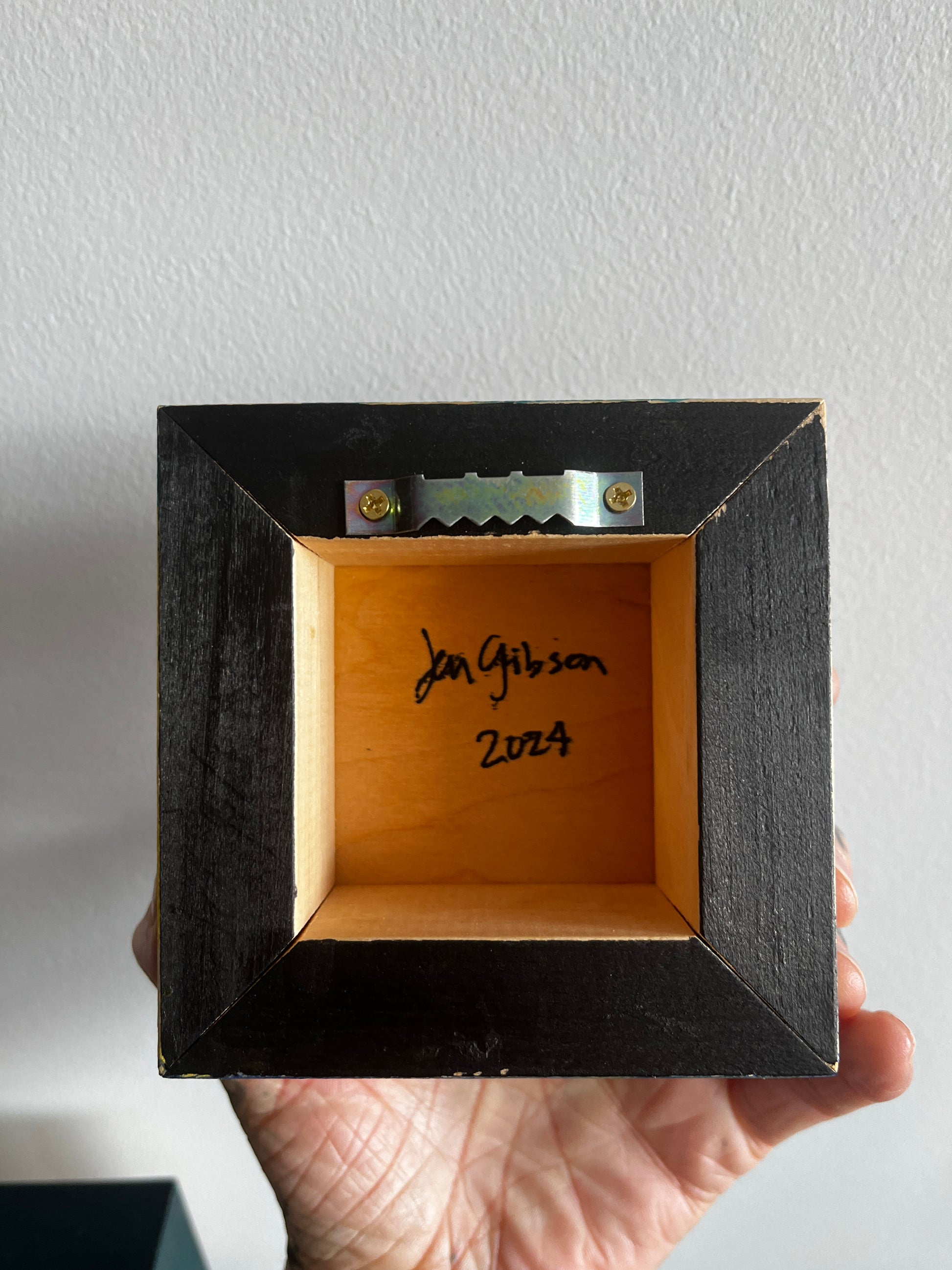 Hand holding a small wooden box with a black frame, signed 'Jan Gibson' and dated '2024'.
