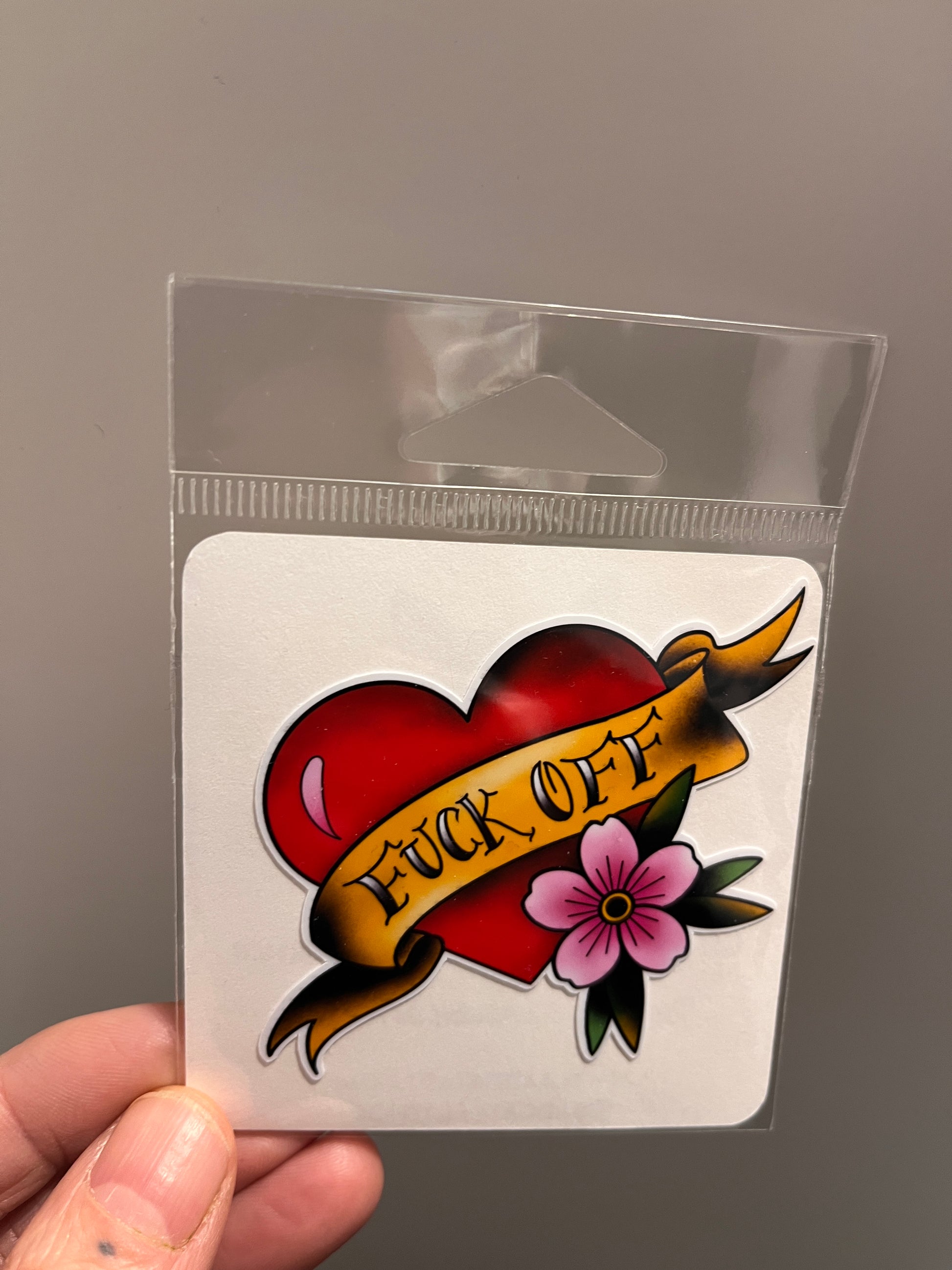 Tattoo-style design with a heart, banner, and flower held in a clear plastic sleeve.