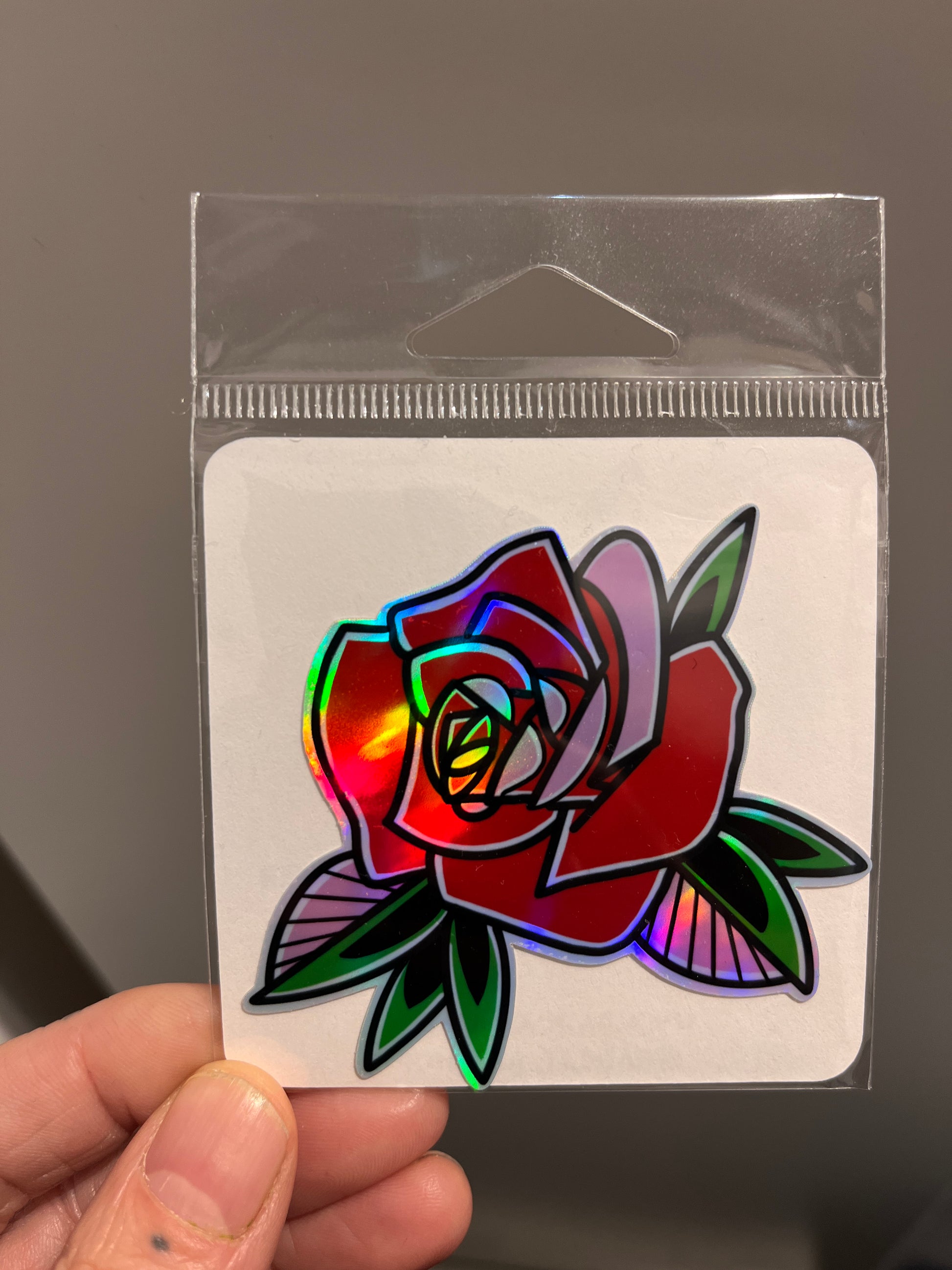 Holographic rose sticker in clear packaging held by a hand against a dark background