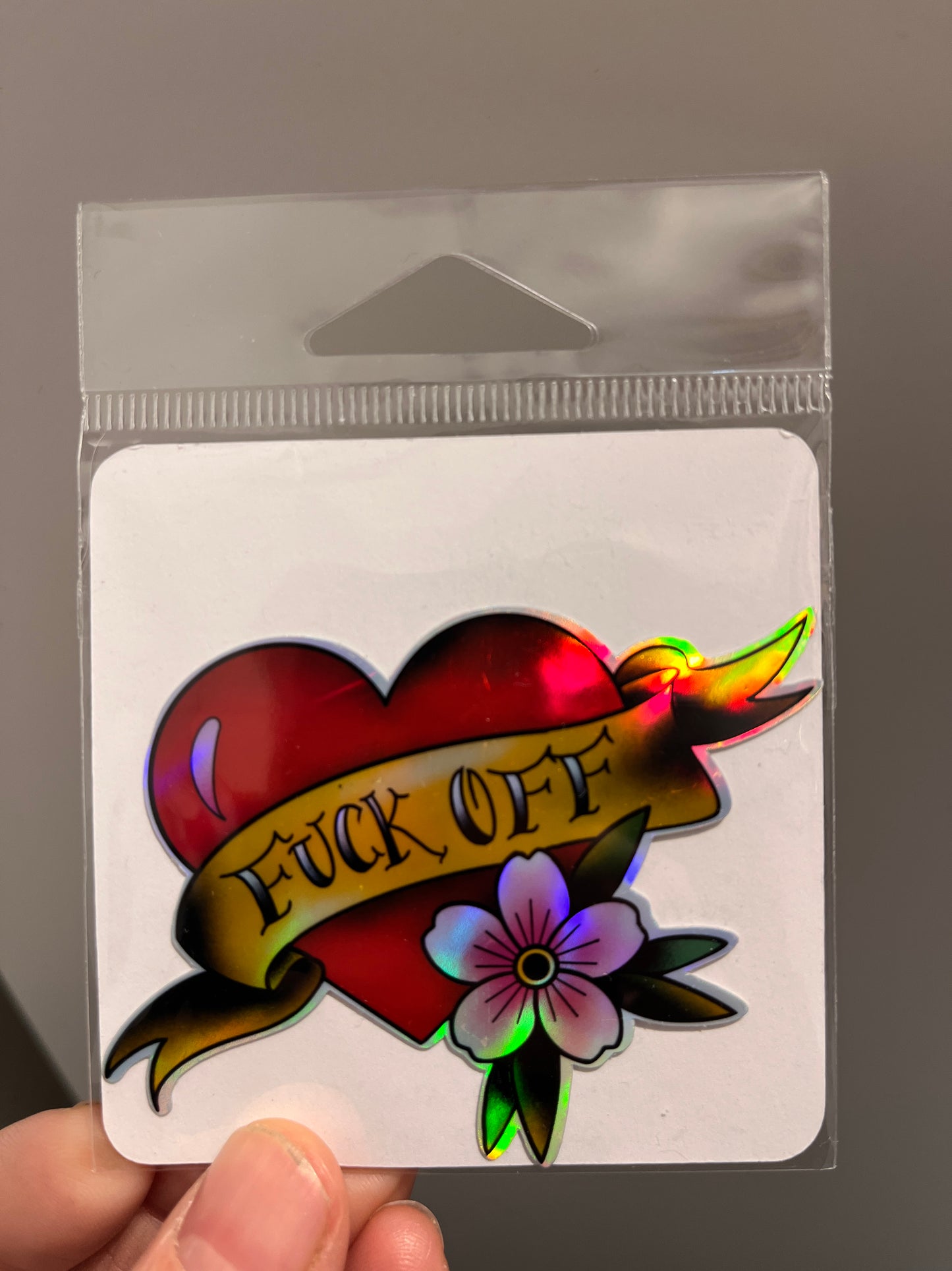 Holographic tattoo design with a heart, banner, and flower held in a clear plastic sleeve.