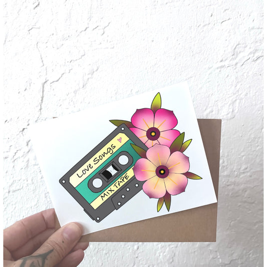 Hand holding a card with a cassette tape and flowers design against a textured white background