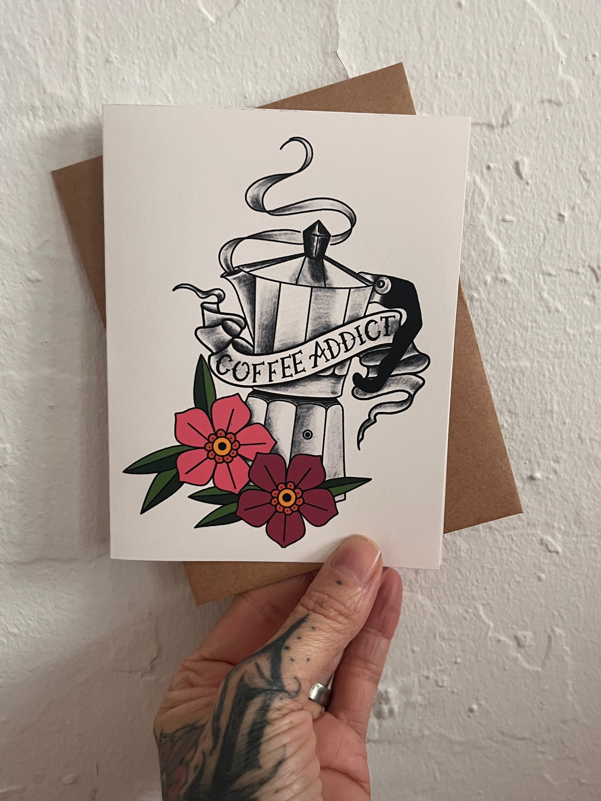 Hand holding a card with tattoo-style design and flowers on a textured white surface