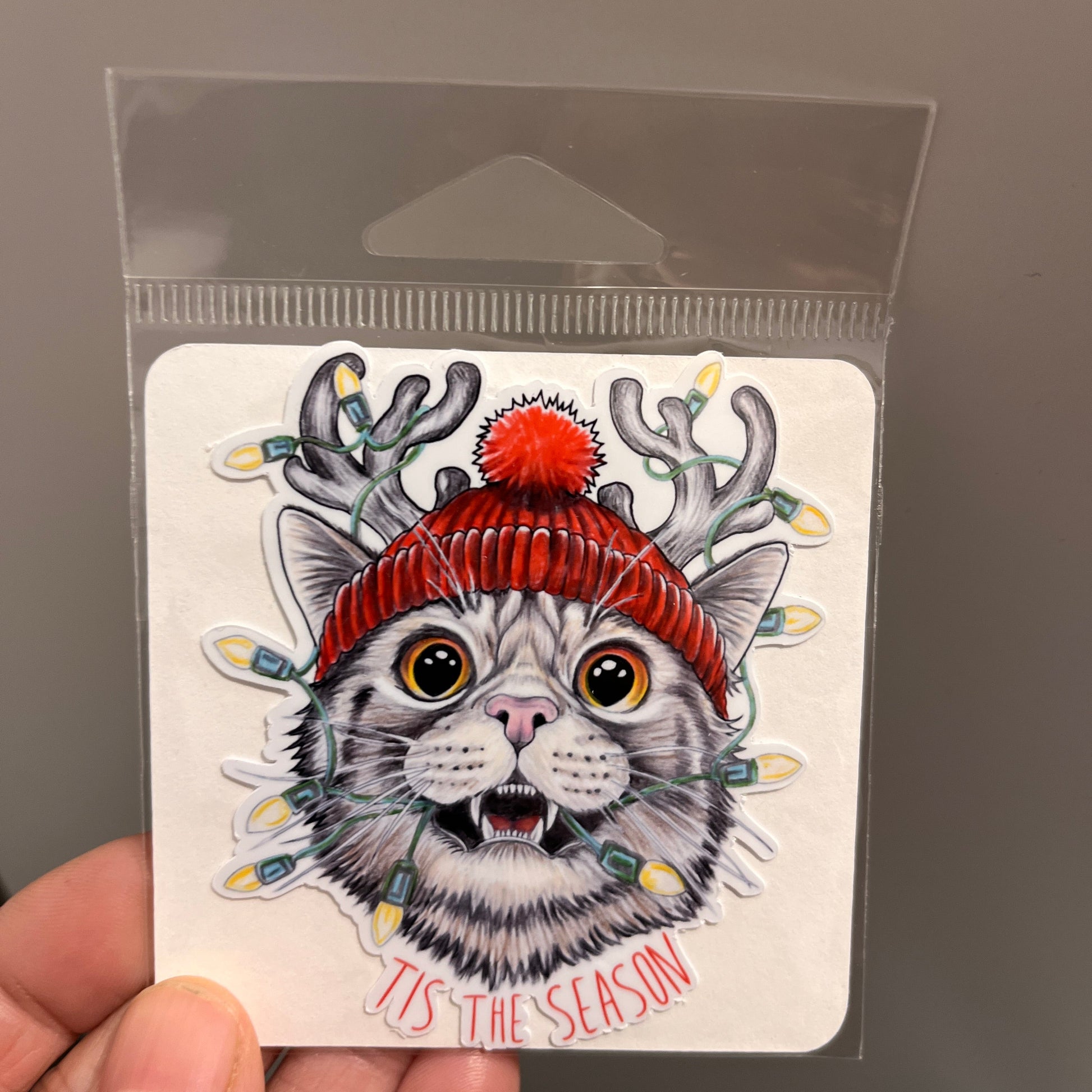 Sticker of a cat wearing a red hat with Christmas lights on a clear card held by a hand.