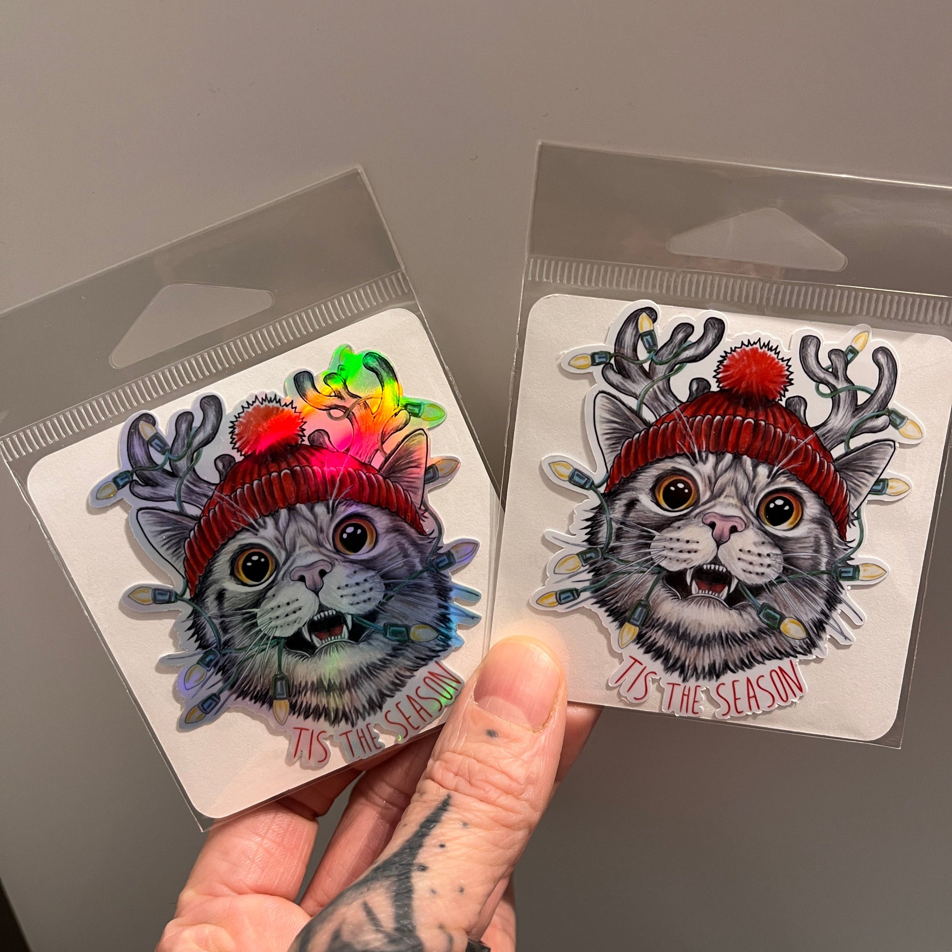 Two colorful cat-themed stickers held by a hand on a gray background