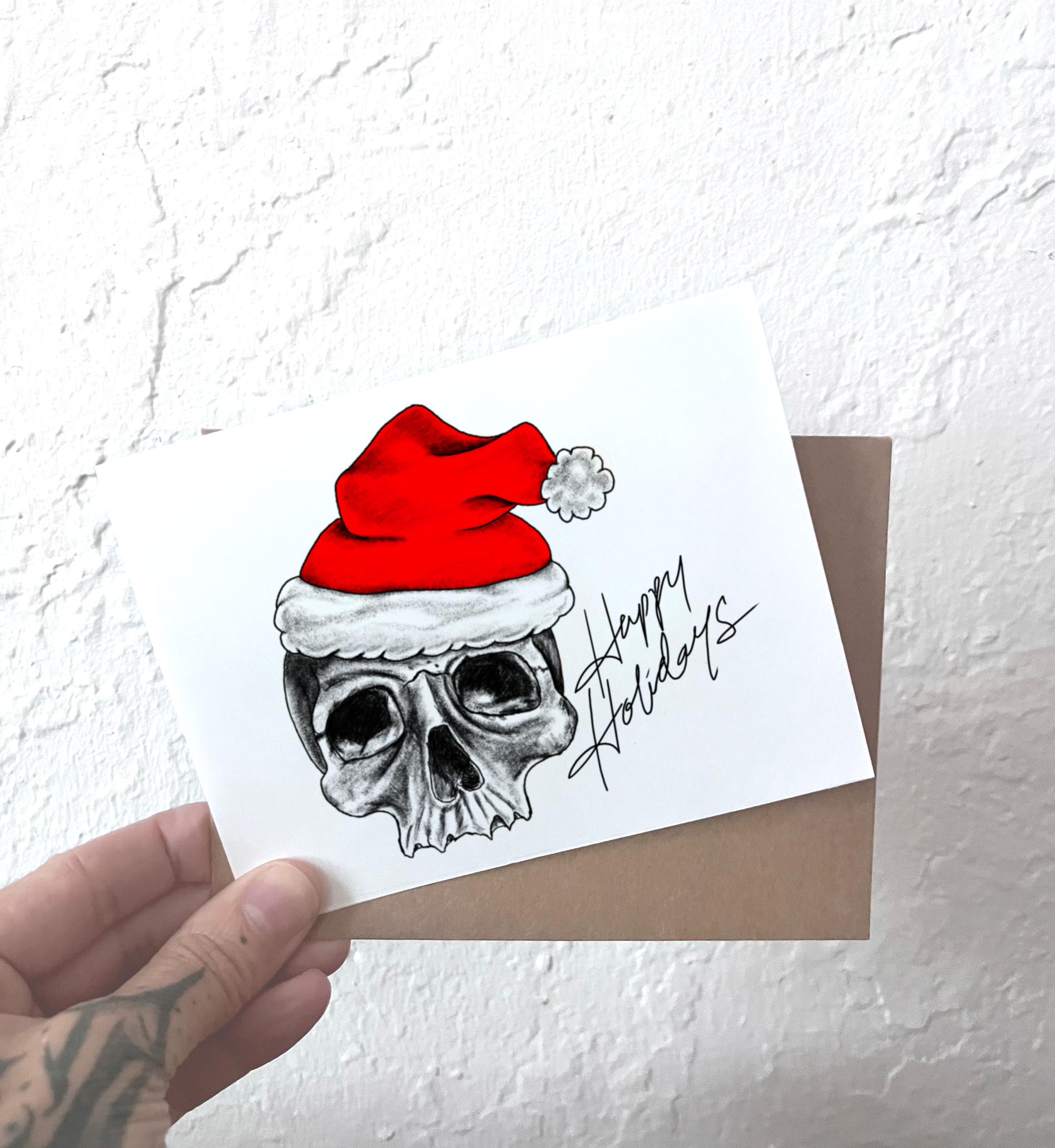Card with a skull wearing a red Santa hat and 'Happy Holidays' text, held against a textured white background.
