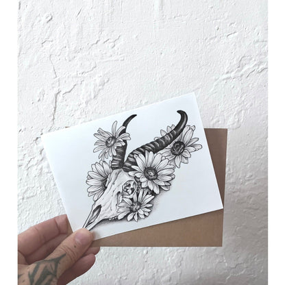 Hand holding a card with a black and white illustration of a deer skull and flowers against a textured white background.
