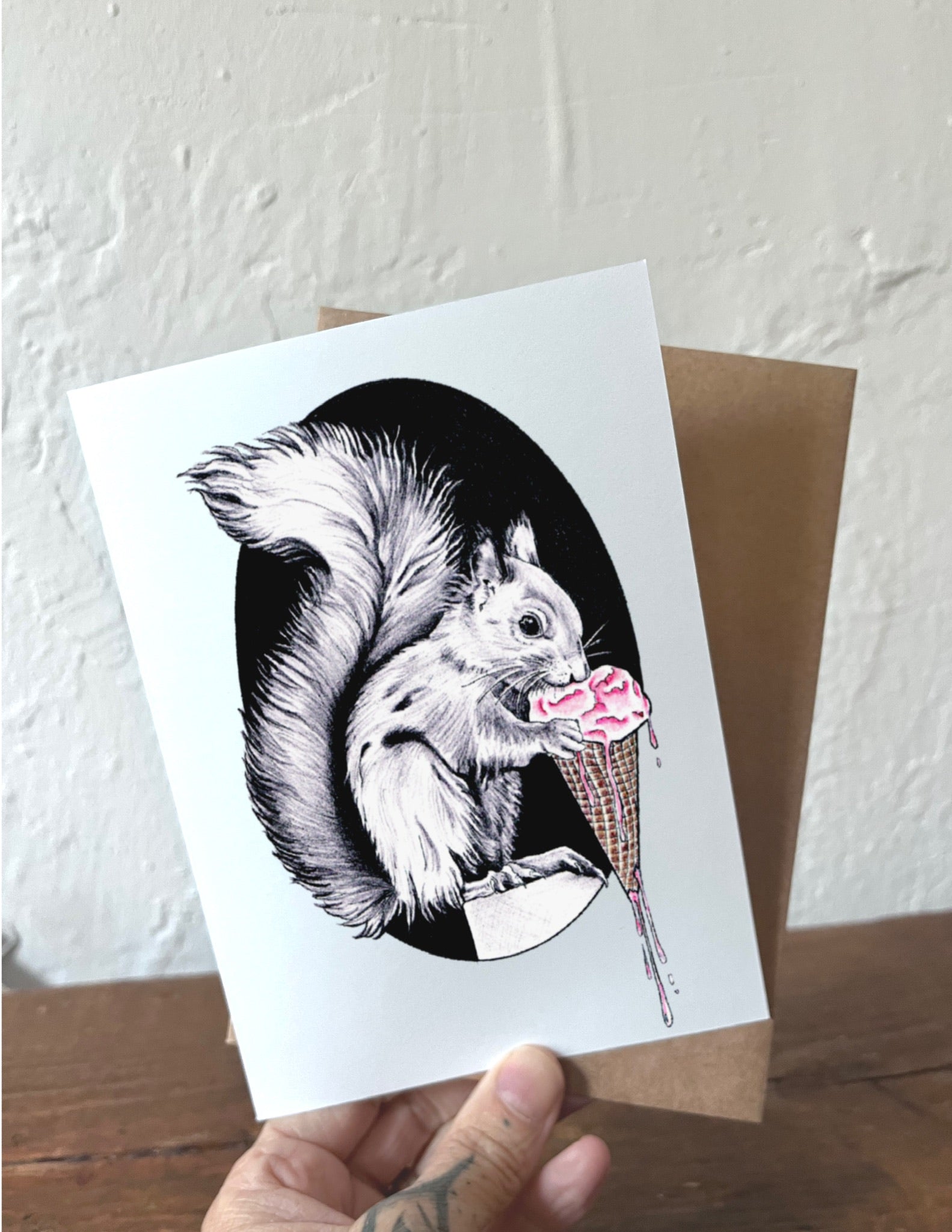 Greeting card with a squirrel eating ice cream illustration held by a hand against a white wall.