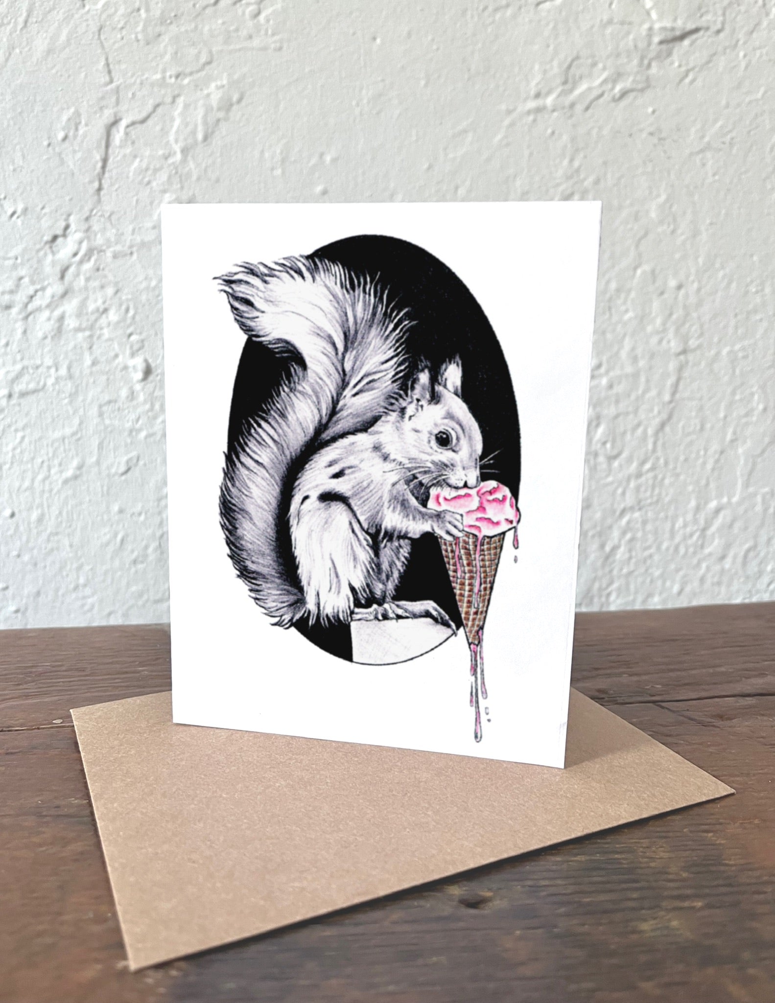 Greeting card with a squirrel and ice cream cone illustration on a white background, placed on a wooden surface with a brown envelope.