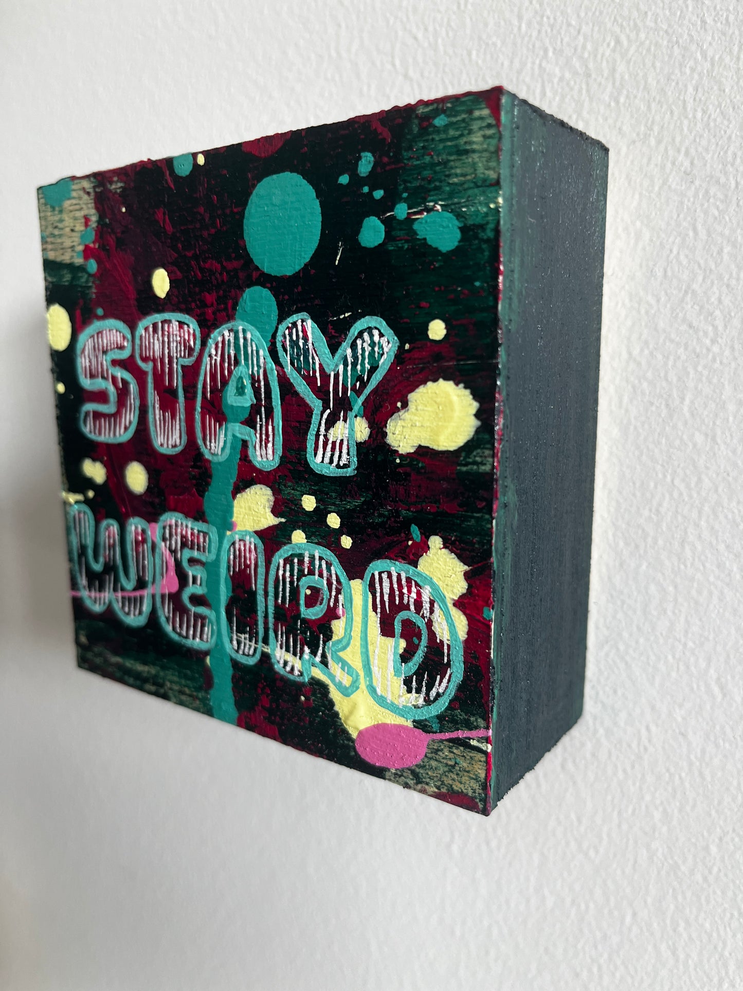 Decorative square wood panel with colourful abstract design and text "Stay Weird" on a white background