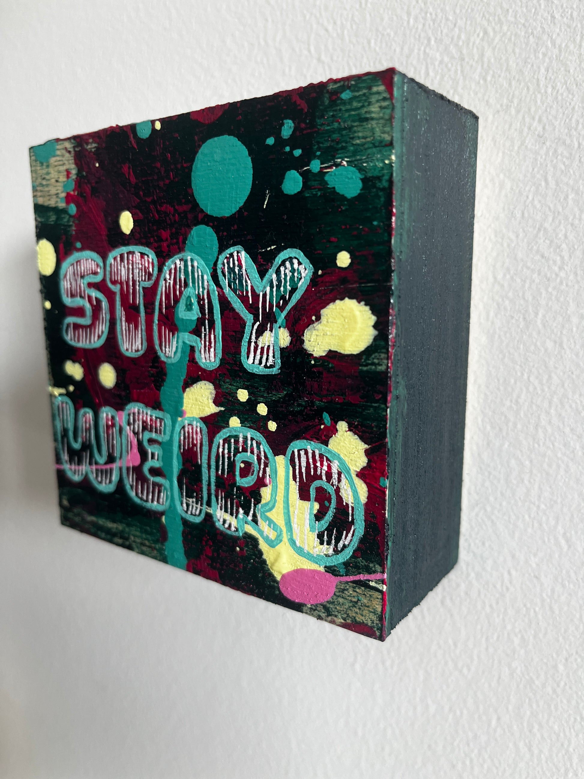 Decorative square wood panel with colourful abstract design and text "Stay Weird" on a white background