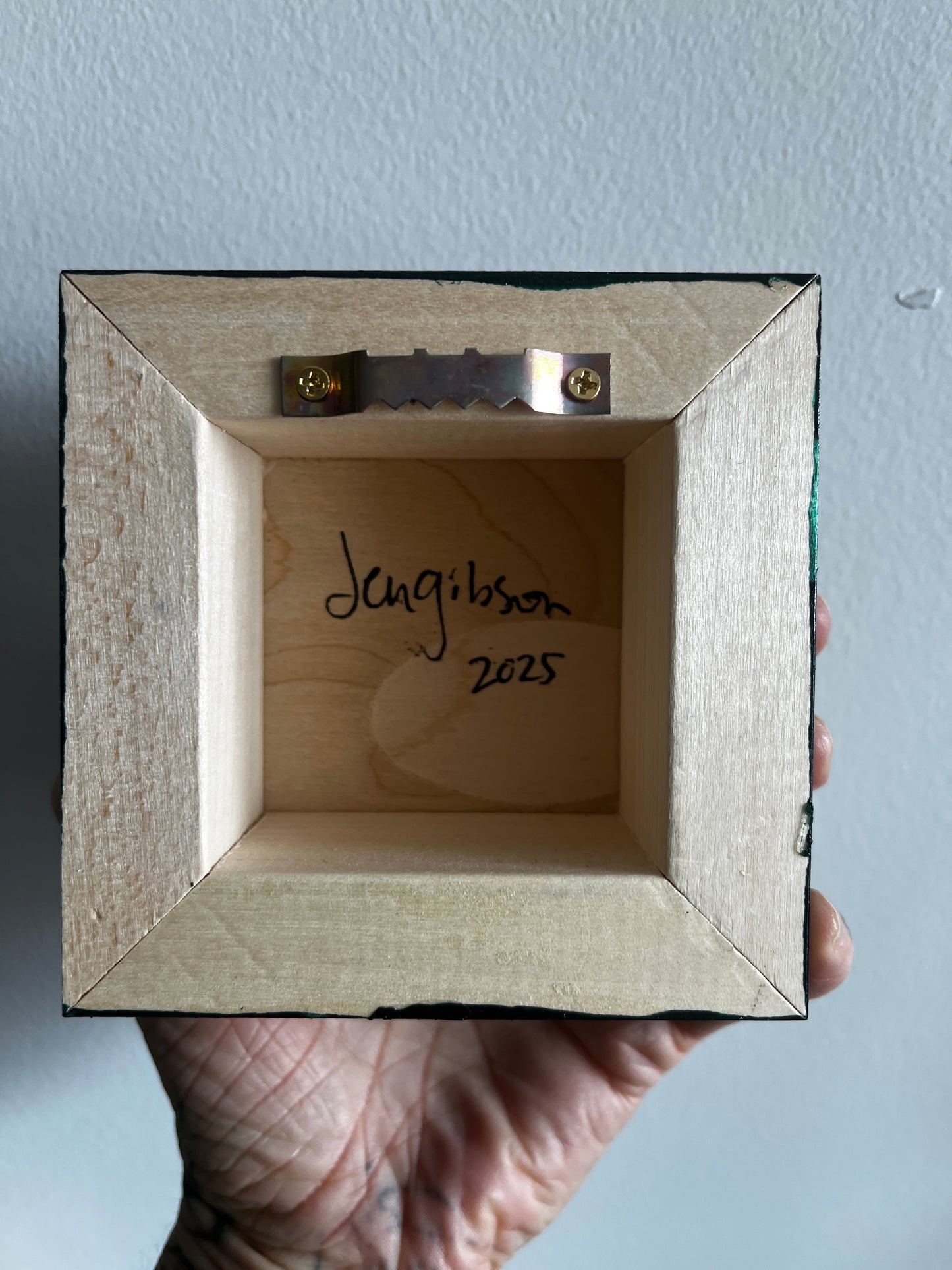 Wooden picture frame with 'Jen Gibson 2025' inscription held by a hand against a light blue background