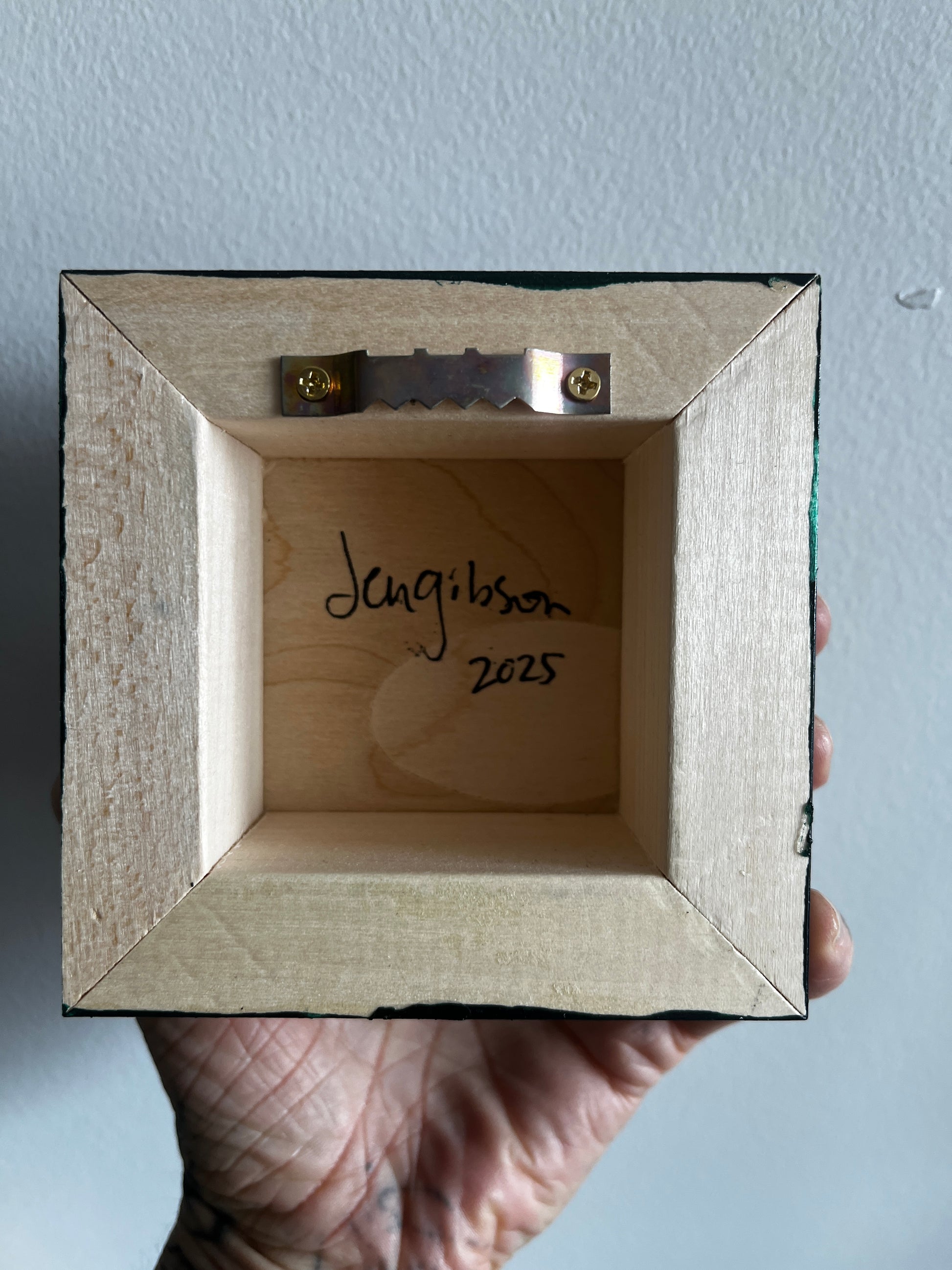Wooden picture frame with 'Jen Gibson 2025' inscription held by a hand against a light blue background