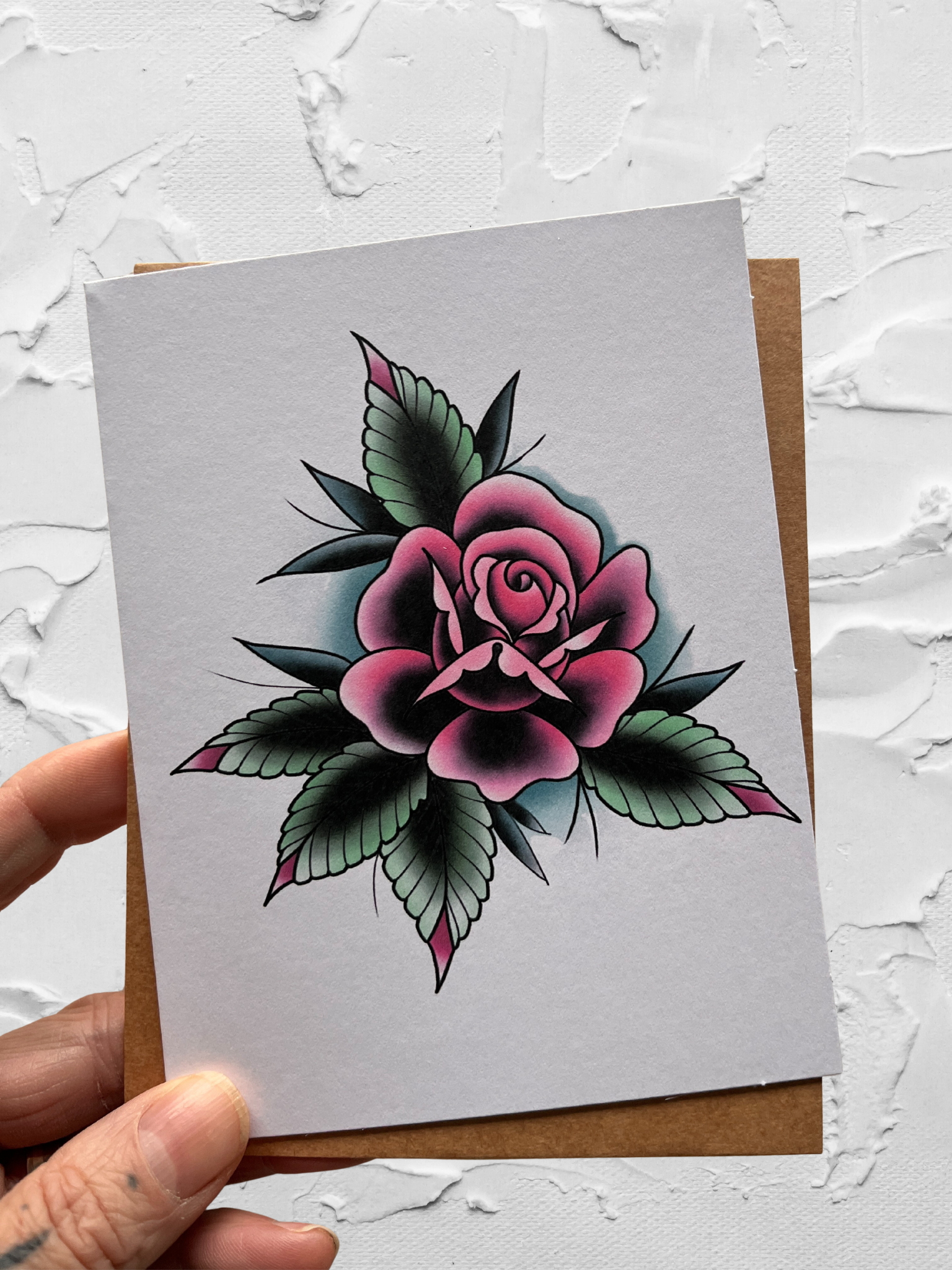 Card with a pink rose design held by a hand on a textured white background