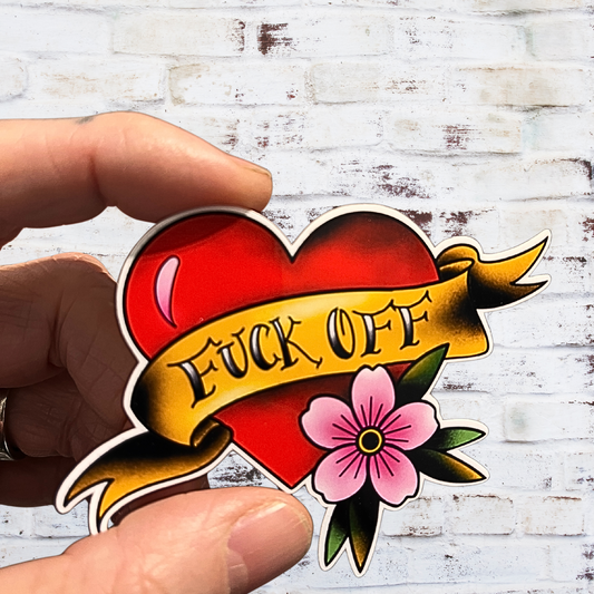 Heart-shaped tattoo design with 'FUCK OFF' text and a flower, held against a white wooden background.