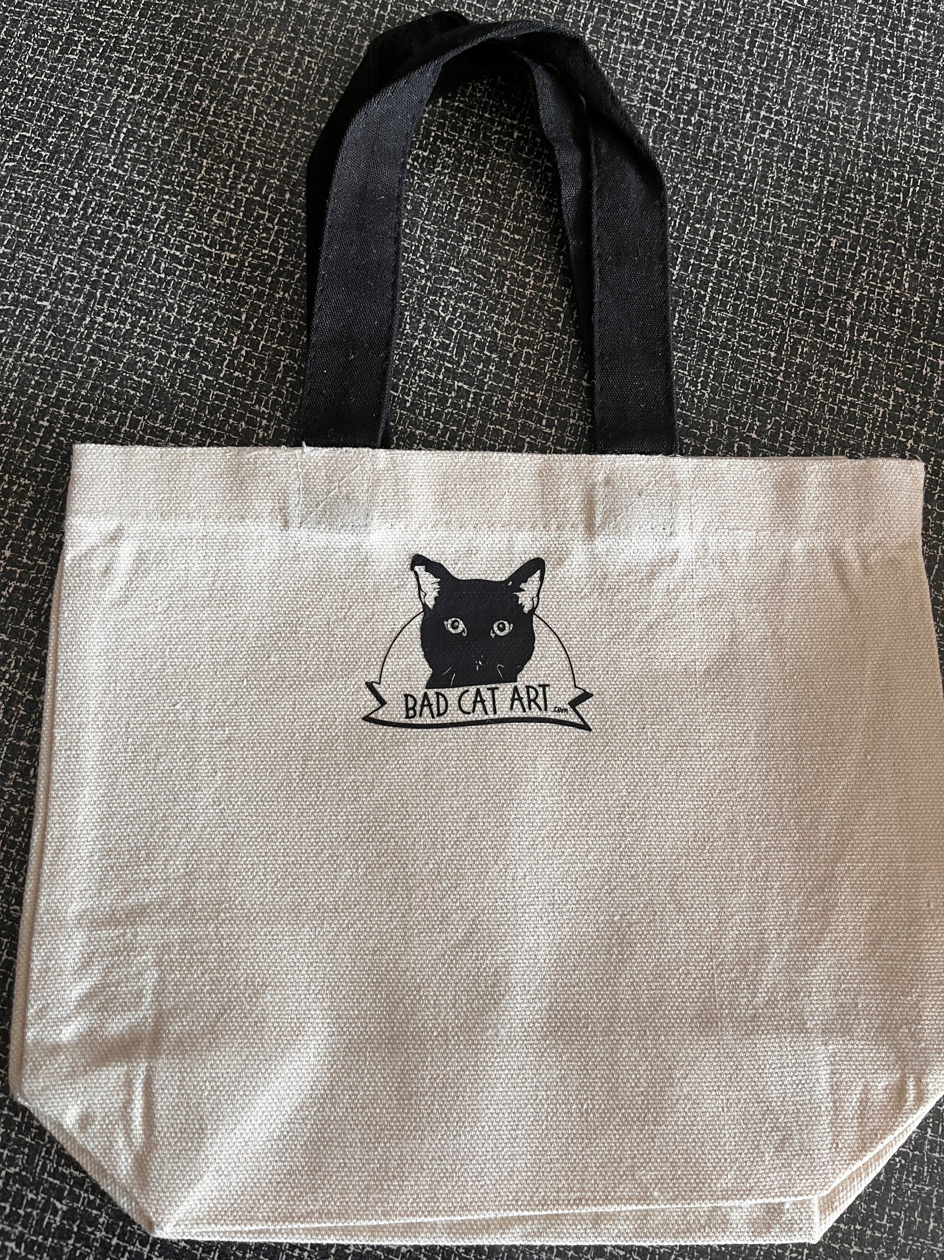 White tote bag with black cat illustration and 'Bad Cat Art' text on a textured surface