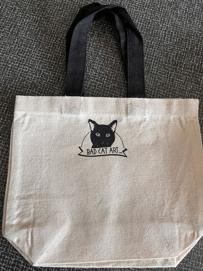 White tote bag with black cat illustration and 'Bad Cat Art' text on a textured surface