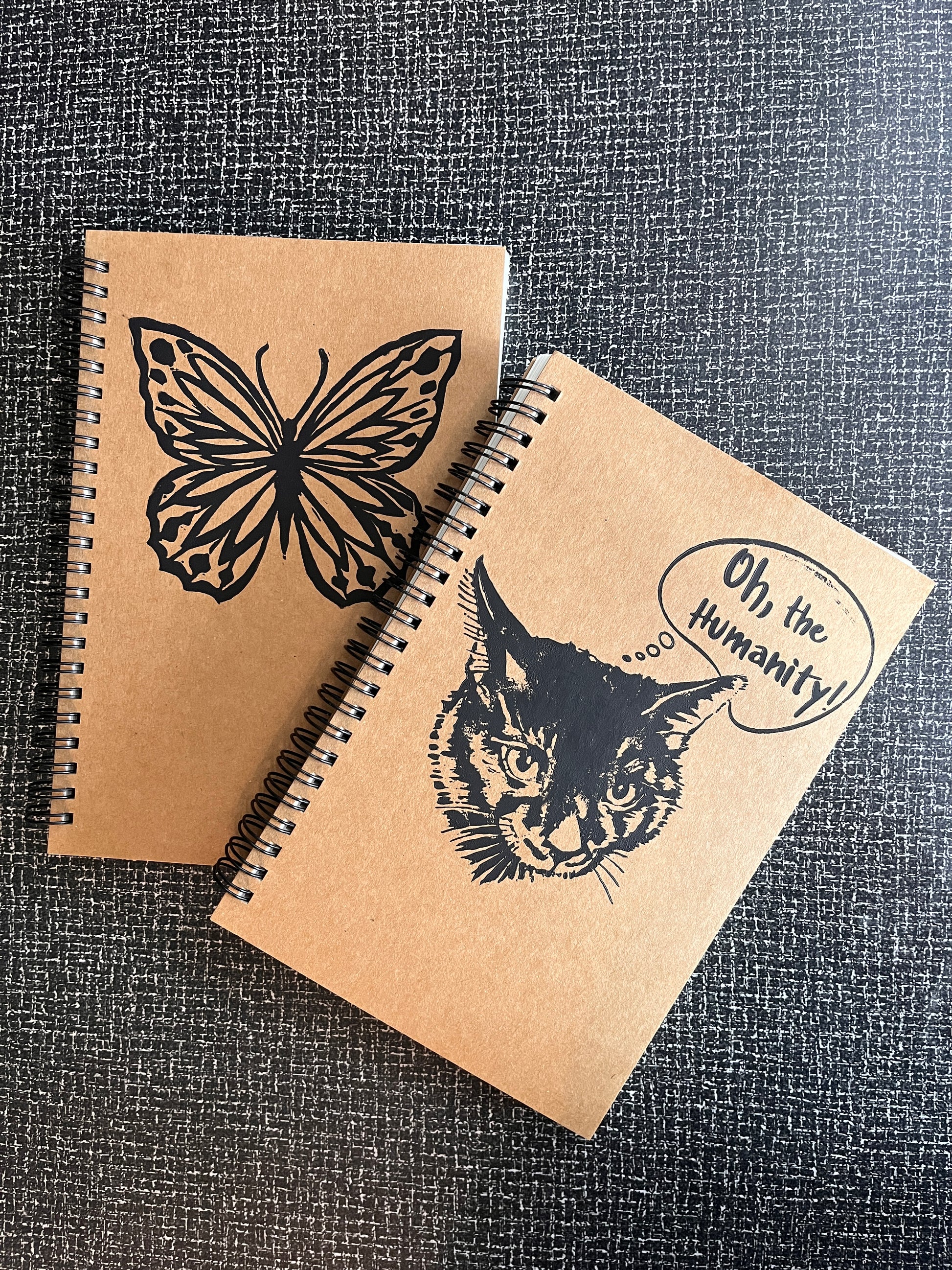 Two brown notebooks with black butterfly and cat illustrations on a textured gray background