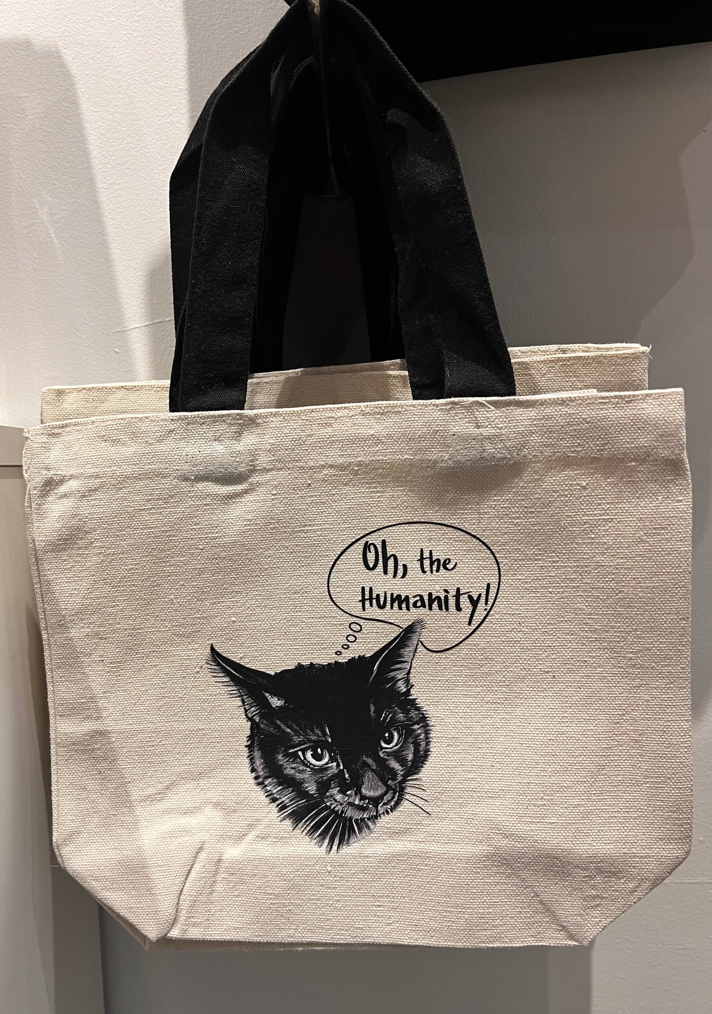 Beige tote bag with black cat illustration and 'Oh, the Humanity!' text on a white background