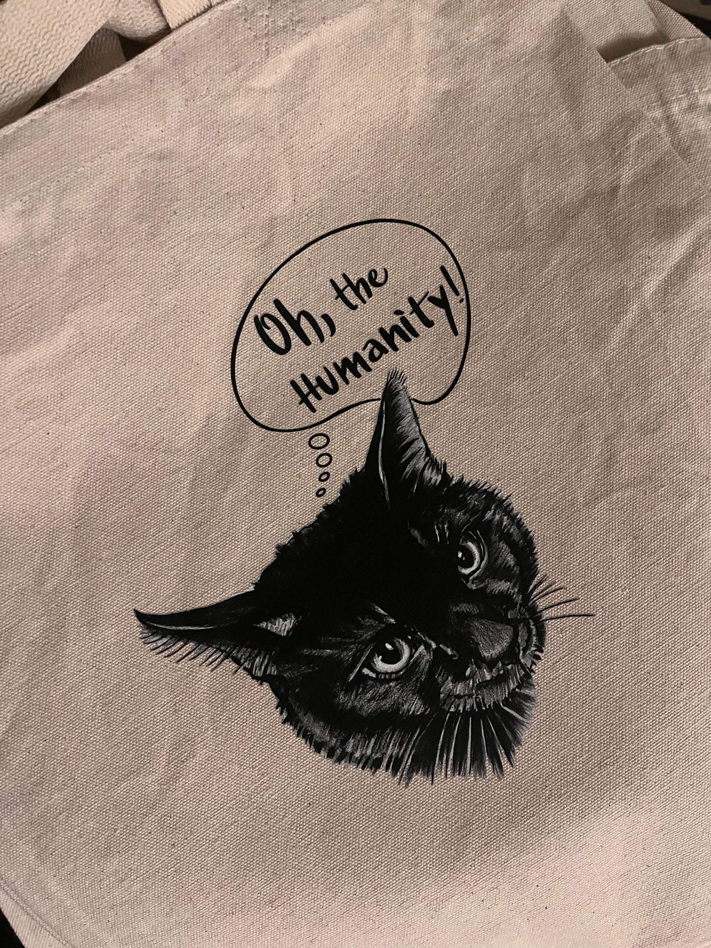 close up view of a neutral canvas tote bag with a black cat and a thought bubble saying oh, the Humanity
