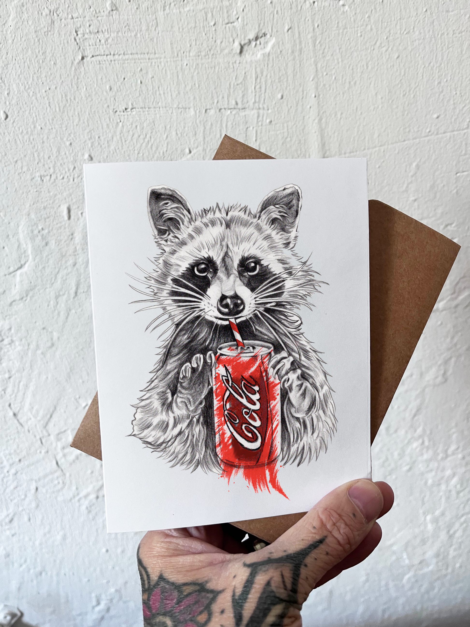 raccoon sipping on a can of cola greeting card being held by a hand in front of a textured white background