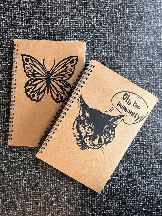Two brown notebooks with black illustrations of a cat and a butterfly on a textured grey surface.