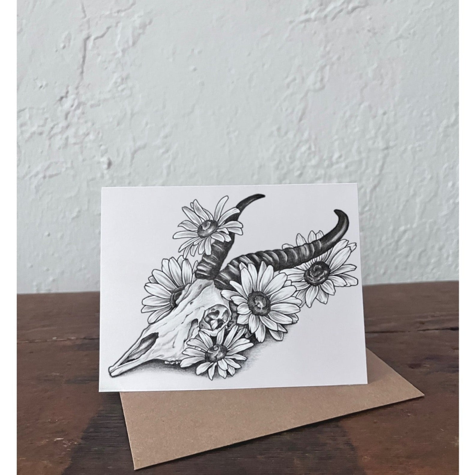 Greeting card with black and white illustration of a springbok skull and daisy flowers on a wooden surface.