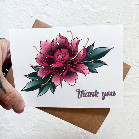 Card with pink floral design and 'Thank you' text held by a tattooed hand on a textured white background.