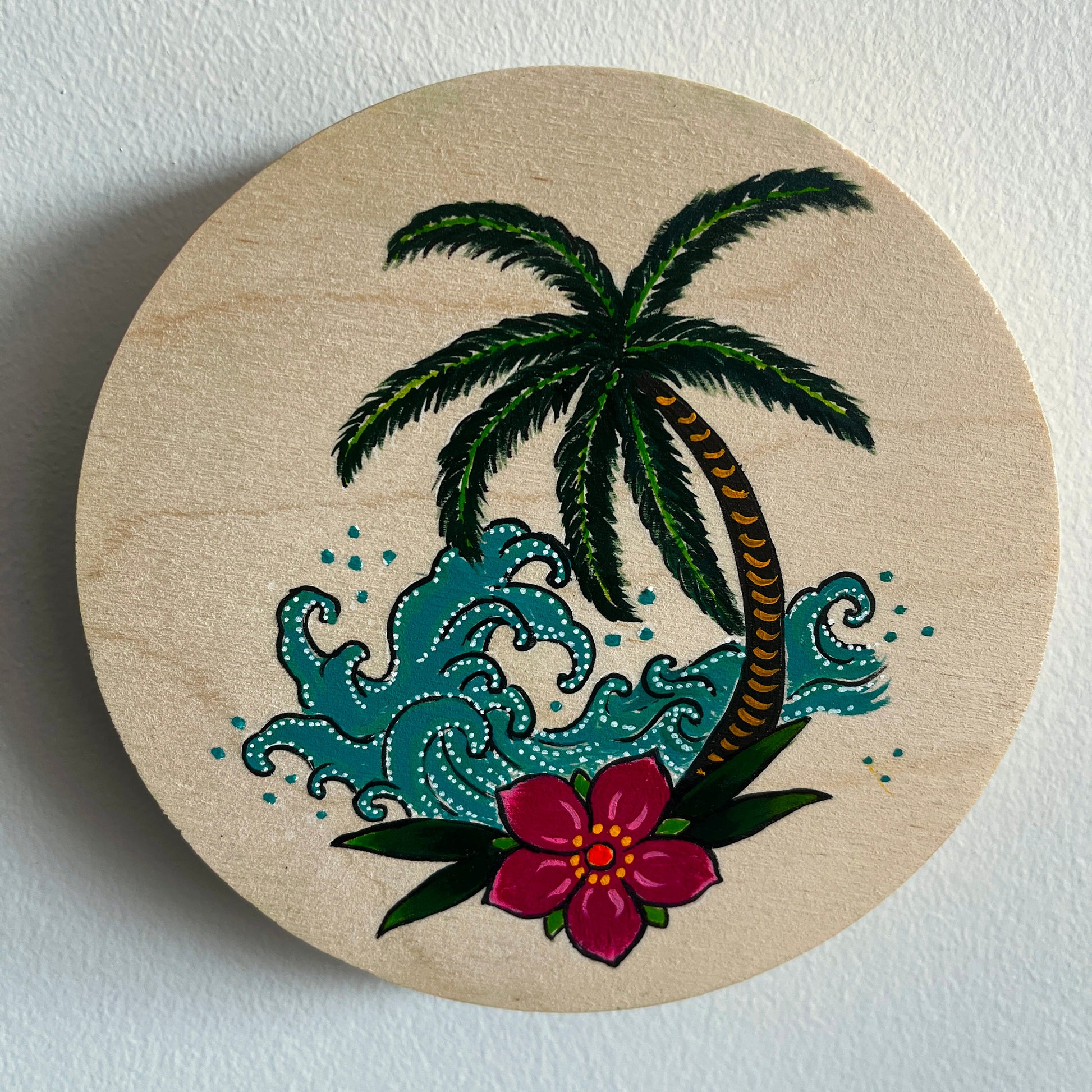 Circular wooden art panel with a hand painted tropical design featuring a palm tree, flowers, and waves on a white background.