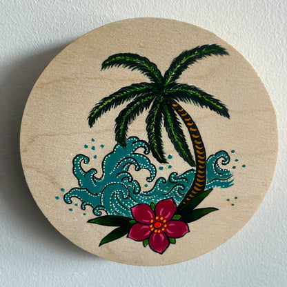 Circular wooden art panel with a hand painted tropical design featuring a palm tree, flowers, and waves on a white background.