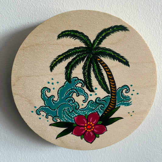 Circular wooden art panel with a hand painted tropical design featuring a palm tree, flowers, and waves on a white background.