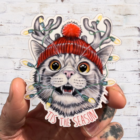 Sticker of a cat wearing a red hat with antlers and Christmas lights, held against a white wooden background.